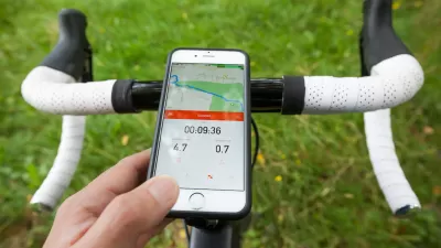 Close-up of bike handlebars with smart phone showing Strava app in handlebar holder. Green grass in background.