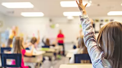 View from back of classroom of young children at desks raising hands.