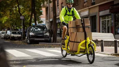 Man in bright yellow jacket riding yellow delivery cargo bike with several brown box packages tied down to bike on urban street.