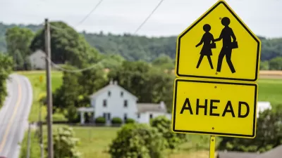 Close-up of yellow and black 'Pedestrians Ahead' sign with blurred farmhouse and road in background.