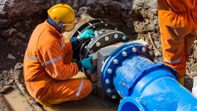 Worker in orange suit and hard hat working on large blue water pipe in ground.