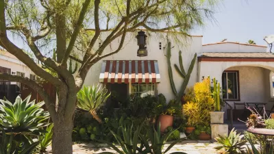 Front of an Spanish style bungalow with striped window awnings and a tree and yard landscaped with cacti.