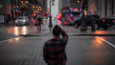 View from behind of person standing on city sidewalk wearing red and black plaid shirt looking across street at dusk.