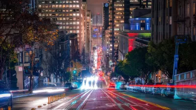 Blurred lights of traffic in motion on San Francisco street with high-rise buildings and Bay Bridge visible in background at night.