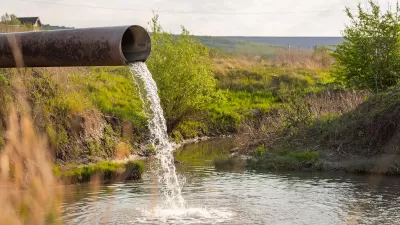 Large metal pipe discharging water or wastewater into creek or body of water with shrubs and field in background.