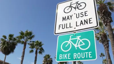 Close-up on white "Bikes may use full lane" and green "Bike route" signs with tall California palm trees in background.
