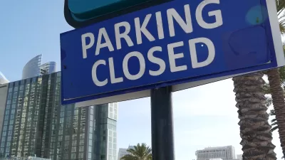 Close-up of blue sign with white text reading PARKING CLOSED with urban glass buildings in background.