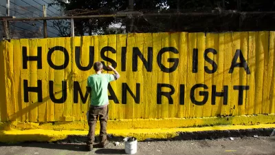 Man painting "HOUSING IS A HUMAN RIGHT" mural in black paint on bright yellow fence