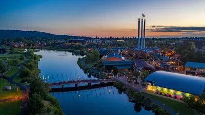 Aerial view of Bend, Oregon with river in foreground and factory smokestacks with American flags on top against sunset sky.