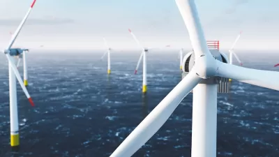 Close-up of top of white offshore wind turbine with other turbines in sea in background.