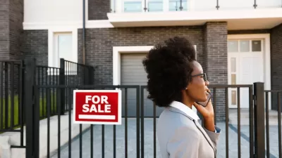 Woman realtor or home buyer client wearing blazer walking past urban condo with red and white For Sale sign while talking on phone.