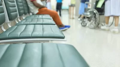 Close-up of blurred empty vinyl chairs with person sitting in background and wheelchair visible in hospital waiting room.