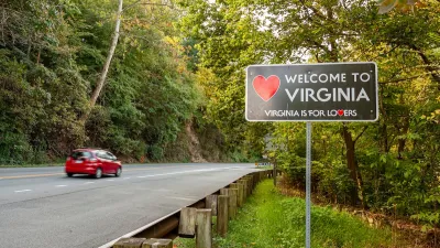 Small red car driving on forested road passing "Welcome to Virginia" sign.
