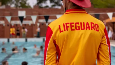 View from behind lifeguard standing in front of crowded public swimming pool wearing yellow shirt with LIFEGUARD in red lettering and red sun hat