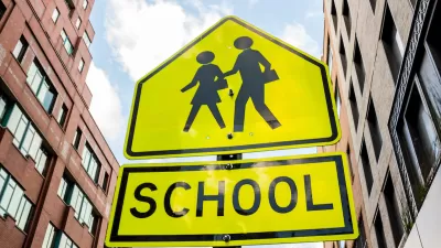 Yellow school crossing sign with two figures and brick buildings in background.