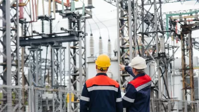 Two men in white and yellow hard hats and matching jackets looking up at power infrastructure in outdoor electrical substation or plant.