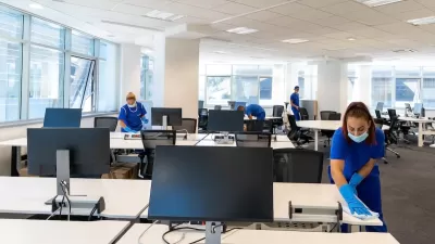 Professional cleaners wearing medical masks and gloves cleaning desks in large empty office.