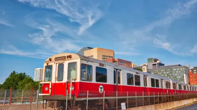 Red T train passing on elevated rail with Boston buildings in background.
