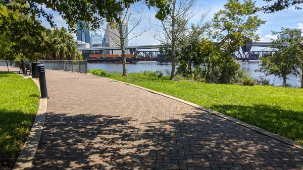 Brick path through an urban park shaded by trees with a river on the righhand side and high-rise buildings in the distance.