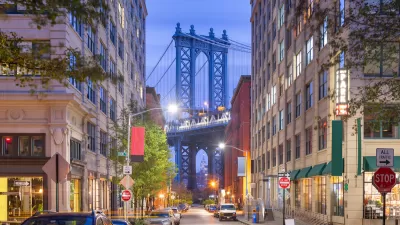 View of Brooklyn Bridge at night from Brooklyn alley with multi-story buildings and parked cars.