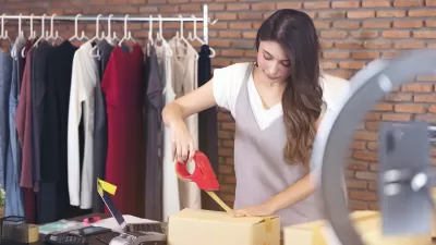 Woman taping up a brown paper package with rack of clothing in background.