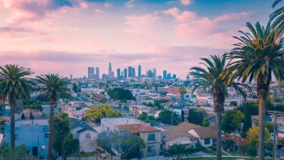 View of downtown Los Angeles from elevated hillside with palm trees in foreground and pink sky.