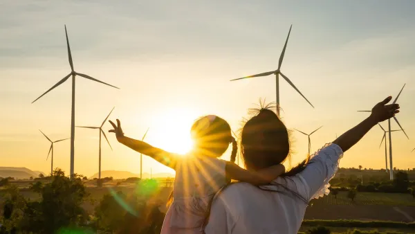 Adult holding young child facing away from camera looking at wind turbines sillhouetted against the sunset.