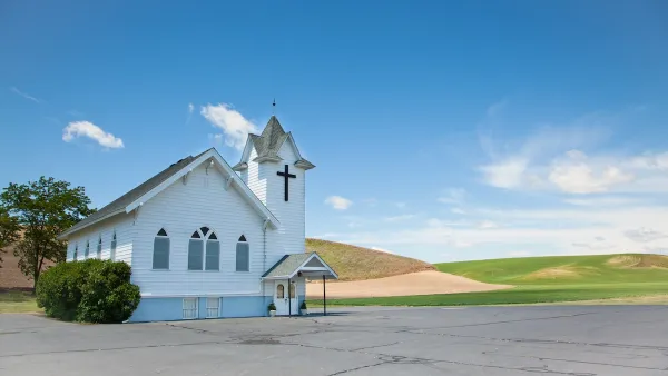 Small white wooden church at edge of large concrete lot with rolling hills or golf course in background.