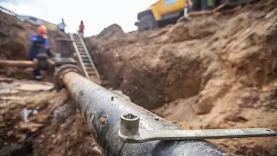 Close-up of exposed pipe in trench with blurred utility workers and yellow truck in background.