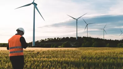 Technician in orange vest and white hard hat standing in field with row of wind turbines.