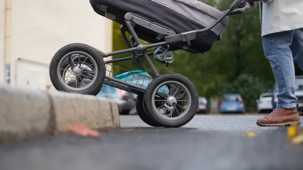 Close-up of bottom half of stroller being pushed onto sidewalk with no curb cut by person in jeans and brown shoes.