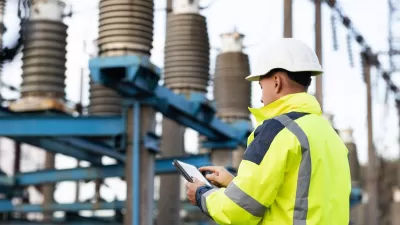Worker in white hard hat and yellow safety jacket holds tablet in front of large electrical utility installation.