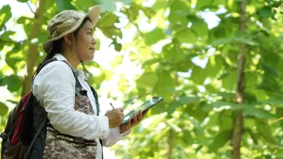 Woman in forest looking at plants and documenting on tablet.