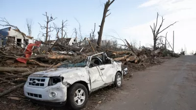 Heavily damaged white small SUV parked on street with heavy tornado damage and leveled houses in Mayfield, Kentucky.