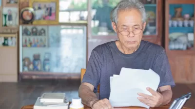 Gray-haired Asian man wearing glasses sitting at wooden table reading documents