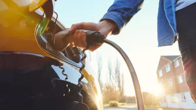 Close-up of hand holding EV charger charging car with brick three-story house in background.