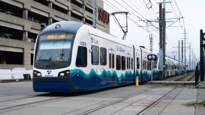 Link Light Rail train travels on ground level tracks paralell to a street with electrical wires overhead.