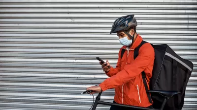 Man in orange jacket wearing surgical mask and bike helmet looks at phone while wearing delivery backpack and holding bicycle