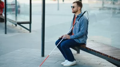 Man with sunglasses and cane sits on bus stop shelter bench.