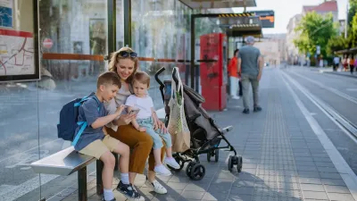 Woman and two children sit on bench at public transit stop waiting for tram with stroller next to them.