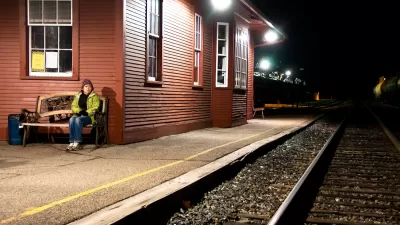 Womanin yellow jacket sitting on bench at outdoor train station with red wood depot building at night.