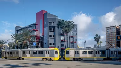 Two silver trains leaving station in Long Beach, California in front of mid-rise residential building.