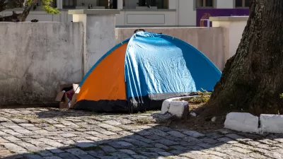 A orange-and-blue tent set up next to a tree in a paved courtyard and a cardboard box of posessions next to the tent entrance.