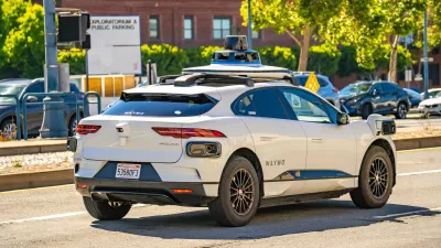 White Waymo self-driving car with roof-mounted camera driving on San Francisco street with Exploratorium parking lot sign in background