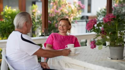 Elderly woman in pink shirt sitting at table on porch across from elderly man in white shirt. A pot of pink flowers sits on the table.