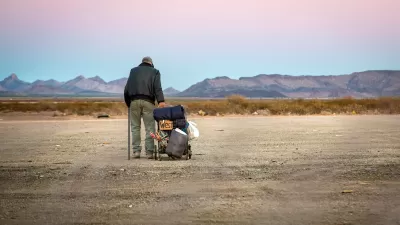 Older unhoused man standing next to cart with belongings and a cardboard sign "West" in a desert landscape.