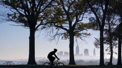 Sillhouette of cyclist riding on path among trees with downtown Cleveland, Ohio skyline visible across water in background.