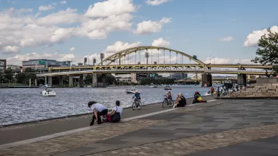 People on bikes and sitting on ledge on Pittsburgh riverfront with arched bridge in background