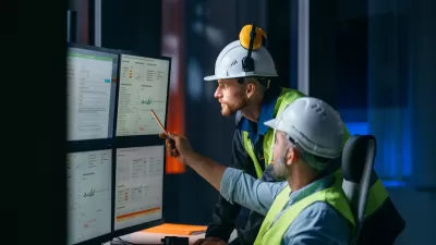 Two workers in yellow safety vests and hard hats looking at multiple computer monitors.