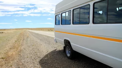 White microbus with yellow stripe on rural road with empty road and blue sky in background.
